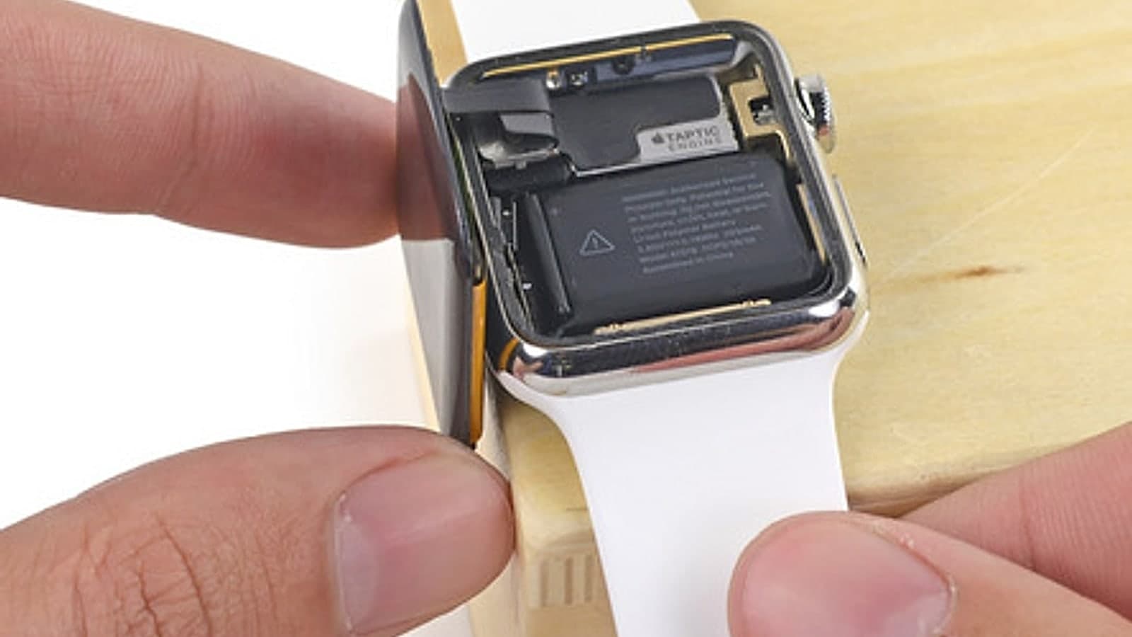 An Apple Watch opened on a bench block, its back off and the battery and Taptic Engine exposed, held steady between two hands.
