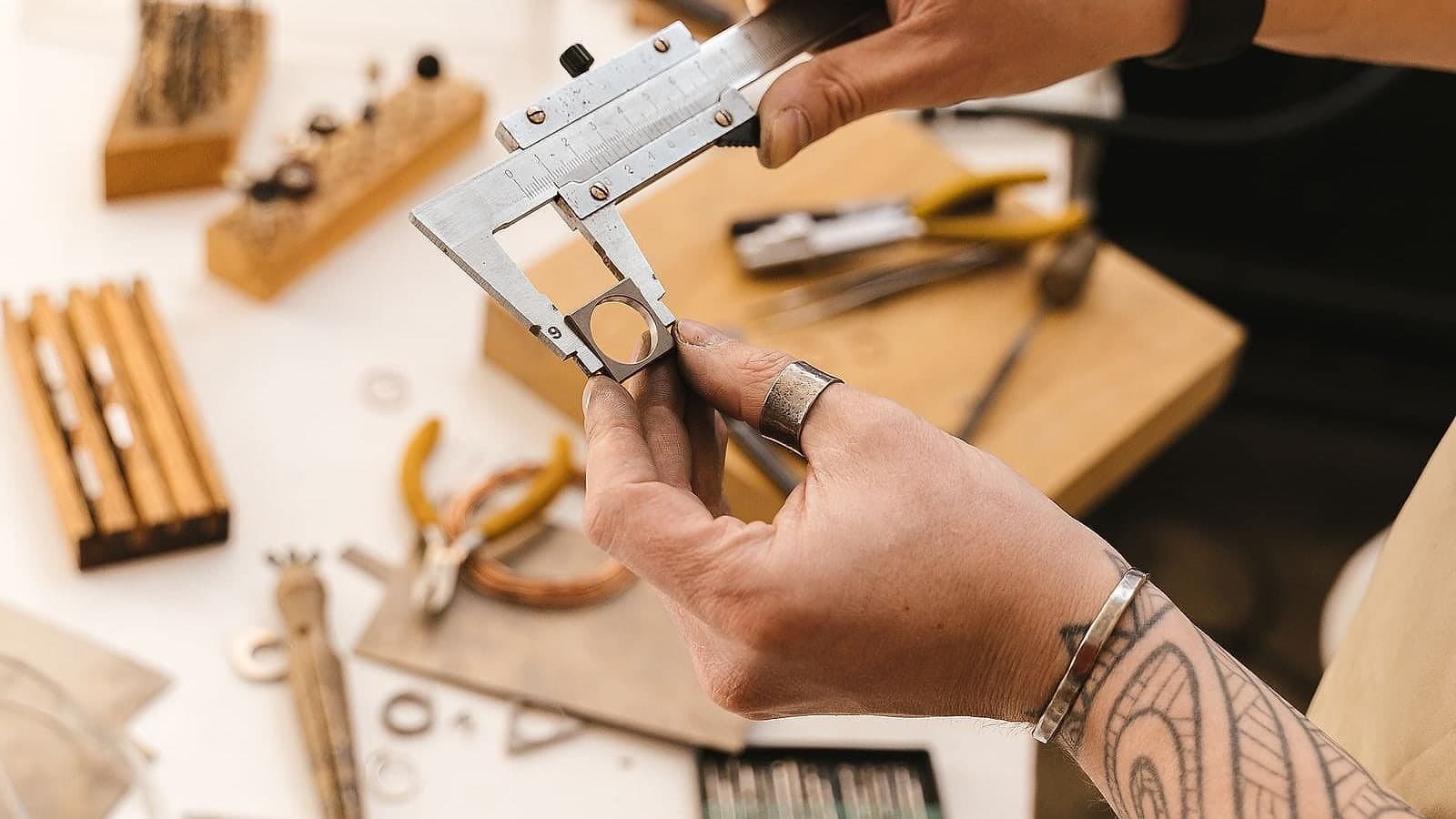 A jeweller measuring a ring with a caliper at a bench laid out with files, pliers and hand tools.